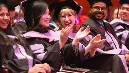 A group of graduates in caps and gowns sit in rows at a commencement ceremony. They are smiling, laughing, and clapping, appearing joyful and celebratory. The lighting is warm, and the focus is on a few graduates in the foreground, including a woman with blonde hair clapping enthusiastically.
