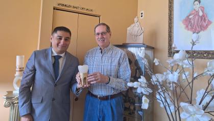 Two men are standing indoors and smiling at the camera. The man on the left is wearing a gray suit and blue tie, while the man on the right is wearing a plaid shirt and blue jeans. They are holding a large model of a tooth together. Behind them is a beige wall, a framed portrait of a young girl in a red dress, a woodcarving of the same girl in the portrait, a display cabinet with figurines, and a branch of white artificial flowers in the foreground.