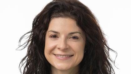 A person with long, curly dark hair is shown wearing a plain black shirt. The individual is positioned against a neutral background.