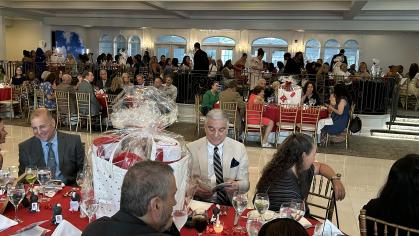 A large banquet hall filled with people seated at round tables covered with red tablecloths. The tables are set with wine glasses, napkins, and small decorations. In the foreground, a group of people is seated around a table with a large wrapped gift in the center. The background shows more guests seated and standing, engaging in conversations. The room has high ceilings and large windows along one wall.