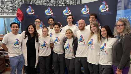 A group of smiling people pose for a photo in front of a "Planet Smilez" banner at a dental outreach event for high school students. Most are wearing matching white t-shirts with the Planet Smilez logo, and a few are in dental scrubs or casual clothing. The setting looks like an indoor community or school space, and there's a colorful mural and balloons in the background.