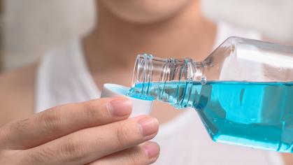 A person is pouring blue mouthwash from a clear plastic bottle into a white cap. The person is wearing a white tank top, and the focus is on the hands and the bottle, with the person's face partially visible in the background.