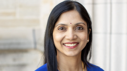 A person with long black hair is wearing a blue blouse and standing in front of a stone wall. 
