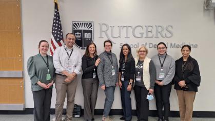 A group of people stand side by side in front of the Rutgers School of Dental Medicine logo and crest, with an American flag positioned to the left.