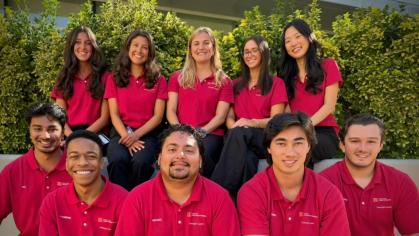 Group photo of ten people wearing matching red polo shirts and dark pants, seated on steps in front of green shrubs. The individuals are arranged in two rows and facing the camera, with sunlight illuminating the scene.