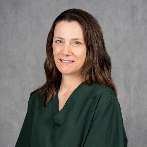 A woman with fair skin and long, straight brown hair parted slightly to the side. She is wearing a dark green scrub top and is smiling gently while facing the camera. The background is a plain gray backdrop.