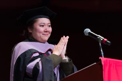 Class president Irene Wong in a graduation cap and gown stands at a podium with a microphone. Her hands are clasped together in front of her chest. The background is dark, and a red cloth is draped over the podium.