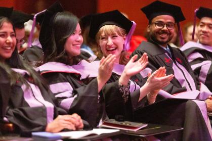 A group of graduates in caps and gowns sit in rows at a commencement ceremony. They are smiling, laughing, and clapping, appearing joyful and celebratory. The lighting is warm, and the focus is on a few graduates in the foreground, including a woman with blonde hair clapping enthusiastically.