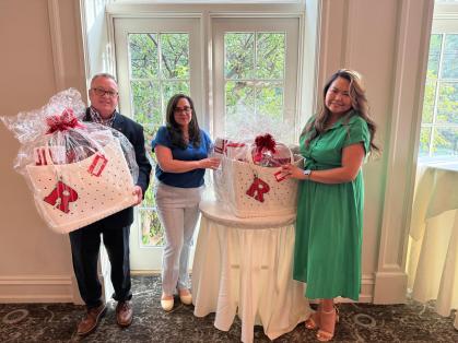 Three people are standing in front of a set of glass doors, each holding or standing next to large white gift baskets wrapped in clear plastic and adorned with red bows. The baskets have a large red letter 'R' on them. The person on the left is wearing a dark suit, the person in the middle is wearing a blue shirt and light pants, and the person on the right is wearing a green dress. The middle basket is placed on a round table covered with a white cloth.