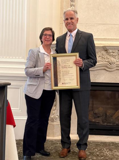 Two individuals are standing side by side in a formal setting, holding a framed certificate together. The person on the left is wearing a striped blazer and dark pants, while the person on the right is dressed in a suit with a light blue tie. The background features an ornate fireplace and elegant wall paneling, suggesting a formal award ceremony or event.