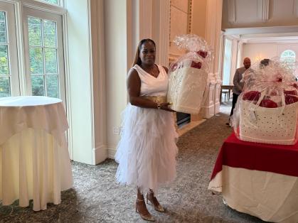 A person wearing a white dress and gold heels is standing indoors, holding a large white basket wrapped in clear plastic with red items inside. The person is near a round table covered with a white cloth and next to another table draped in a red and white cloth, which also holds similar baskets. The room features large windows, light-colored walls, and carpeted flooring.