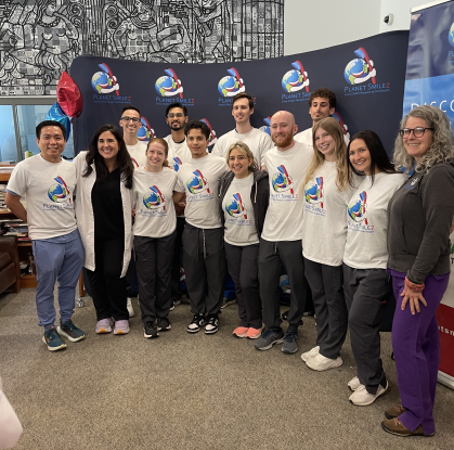 A group of smiling people pose for a photo in front of a "Planet Smilez" banner at a dental outreach event for high school students. Most are wearing matching white t-shirts with the Planet Smilez logo, and a few are in dental scrubs or casual clothing. The setting looks like an indoor community or school space, and there's a colorful mural and balloons in the background.