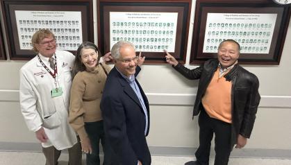 Four individuals standing in a hallway, looking at and pointing toward framed photo collages mounted on the wall. The frames contain rows of black-and-white headshots with text above each collage, indicating they are historical class or alumni photographs. One person is wearing a white lab coat, while the others are dressed in business or casual attire.