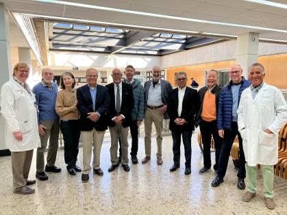 A group of people standing together indoors in a bright, modern space with large windows and skylights. Two individuals are wearing white lab coats, while others are dressed in business or casual attire.