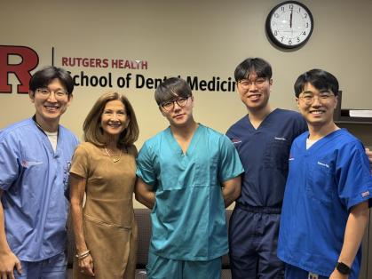 A group of five individuals standing together in front of a beige wall with the Rutgers Health School of Dental Medicine logo. Four people are wearing medical scrubs in shades of blue and teal, while one person is dressed in a brown short-sleeve dress. A clock is visible on the wall above them.