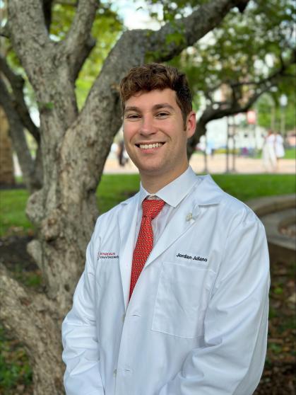 A person stands outdoors in front of a tree, wearing a white coat. They are dressed formally with a red tie and white shirt. 
