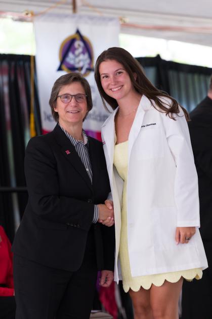 Two individuals are standing and shaking hands at a formal event. One person is wearing a dark suit with a striped shirt, while the other is dressed in a light yellow dress and a white coat.