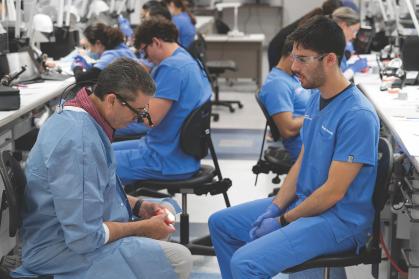 A group of individuals wearing blue scrubs are seated in a clinical laboratory setting. Two people are positioned facing each other in the foreground, one holding a dental model while the other observes.