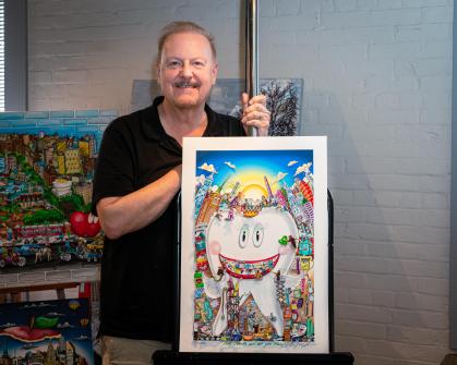 Person wearing a black shirt is holding a colorful artwork featuring a large cartoon-style tooth with a wide smile, surrounded by vibrant cityscape elements, buildings, and playful details under a bright blue sky with clouds and a yellow sun. Additional colorful artworks are displayed in the background against a light gray brick wall.