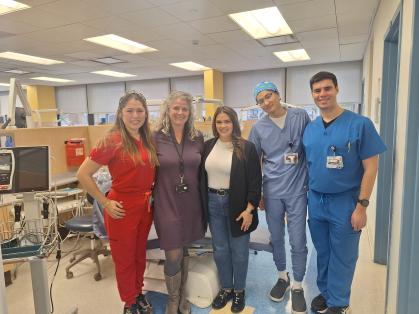Group of five adults standing together in a dental clinic, including several people wearing scrubs and ID badges, posing in front of dental chairs and equipment under bright overhead lights.