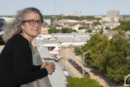 A person with shoulder-length gray hair and round glasses leans on a rooftop railing, looking toward the camera. The person is wearing a dark cardigan and stands against a backdrop of a cityscape with trees, buildings, parked cars, and a road below. The background is softly blurred, drawing focus to the person in the foreground. The photo is taken in daylight under clear skies, with natural light illuminating the scene.