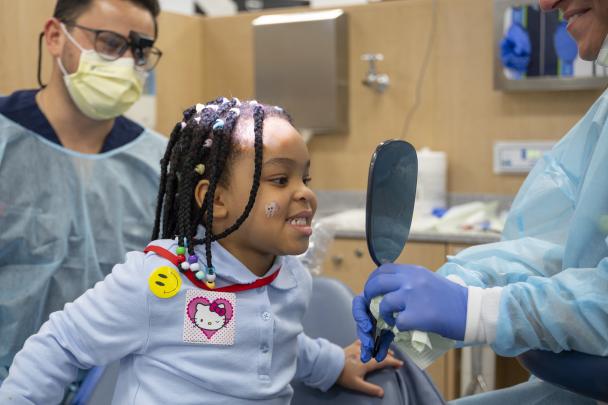 A child sitting in a dental chair looks toward a handheld mirror that a gloved dental professional is holding.